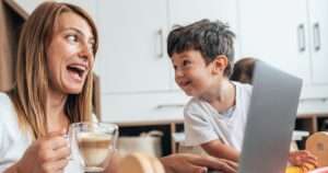 Eine Mama sitzt mit ihrer Tasse Kaffee am Tisch, vor ihr der geöffnete Laptop. Ein Junge sitzt ebenfalls am Tisch, ihr zugewandt und schaut sie fröhlich an, während ihr es schwer fällt, ihre klare Grenzen setzen, weshalb ihr Gesichtsausdruck zeigt, dass sie in Bedrängnis ist.