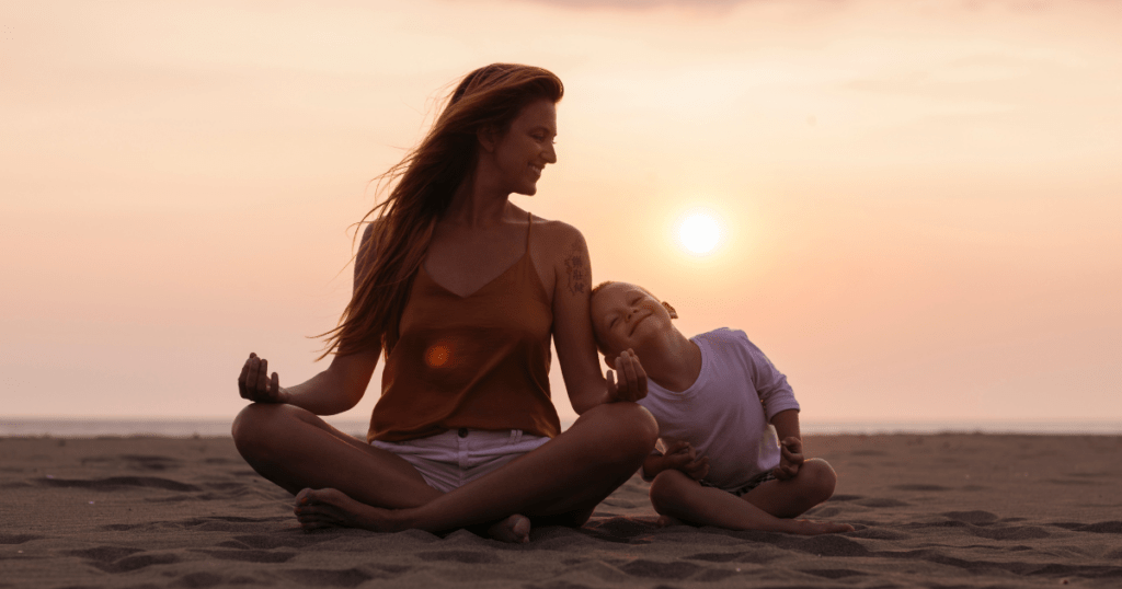 Eine meditierende Mama sitzt am Strang, hinter ihr geht die Sonne unter, sie schaut zur Seite und lächelt ihr Kind an, das neben ihr sitzt.